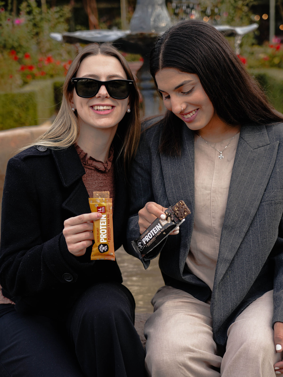 Dos amigas sonriendo al aire libre, sosteniendo Barritas de Proteína Underfive sabor Salted Peanut y Cacao, snack saludable para compartir