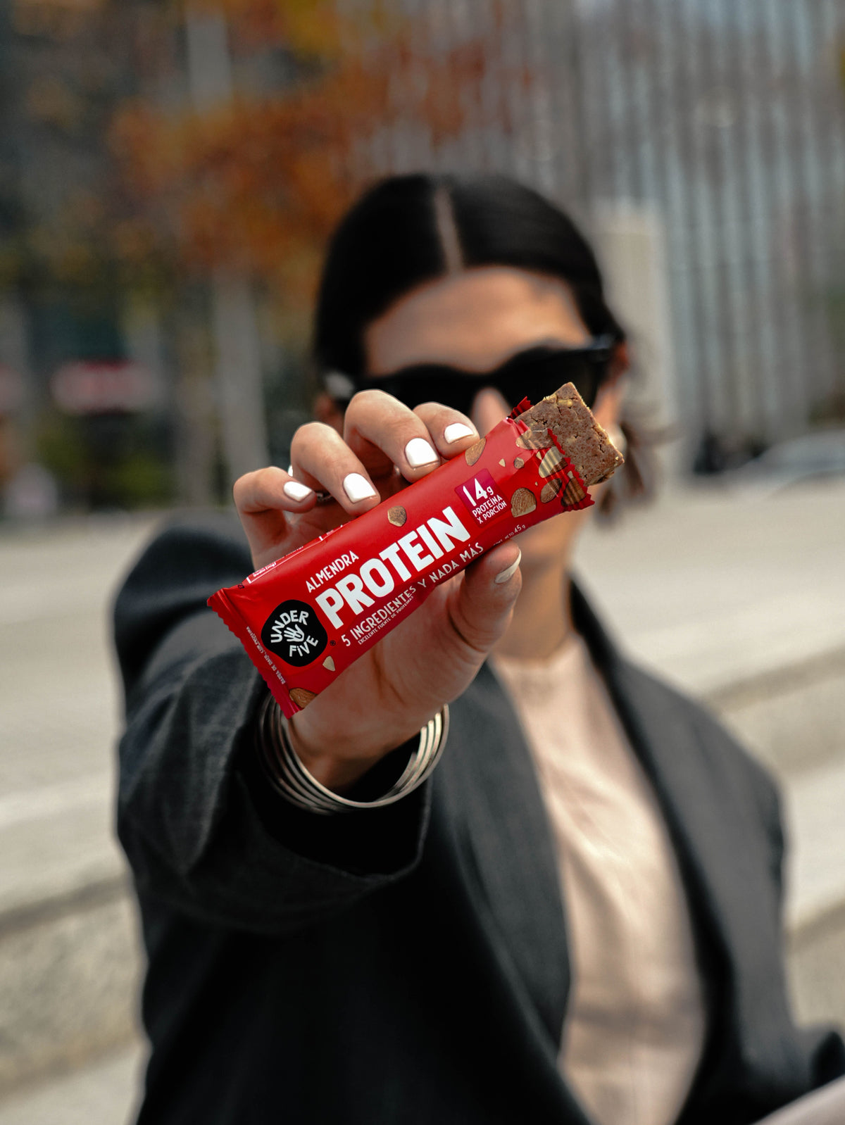 Mujer sosteniendo una Barrita de Proteína Underfive sabor Almendra, 14g de proteína por porción, snack saludable en la calle, contexto urbano