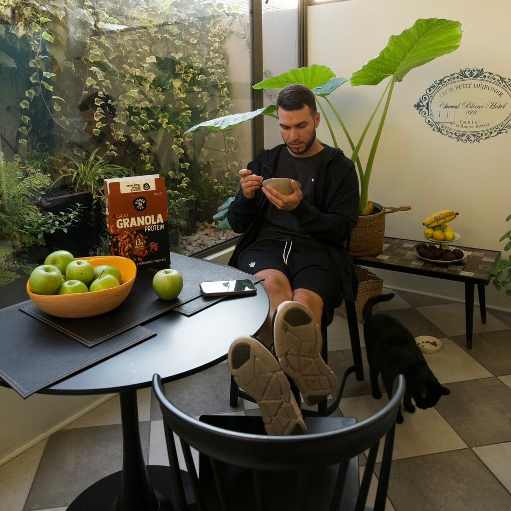 Hombre desayunando en casa un bowl de Granola Protein Underfive sabor Cacao, en una mesa con manzanas verdes, junto a una planta y un gato negro