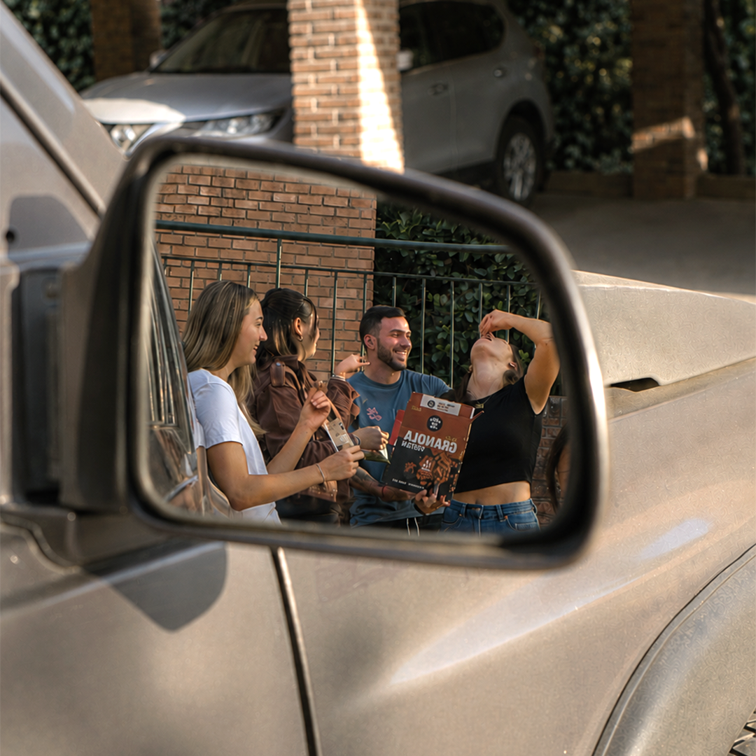 Reflejo en espejo retrovisor de auto: grupo de amigos compartiendo Granola Protein Underfive al aire libre, una persona comiéndola directo de la caja